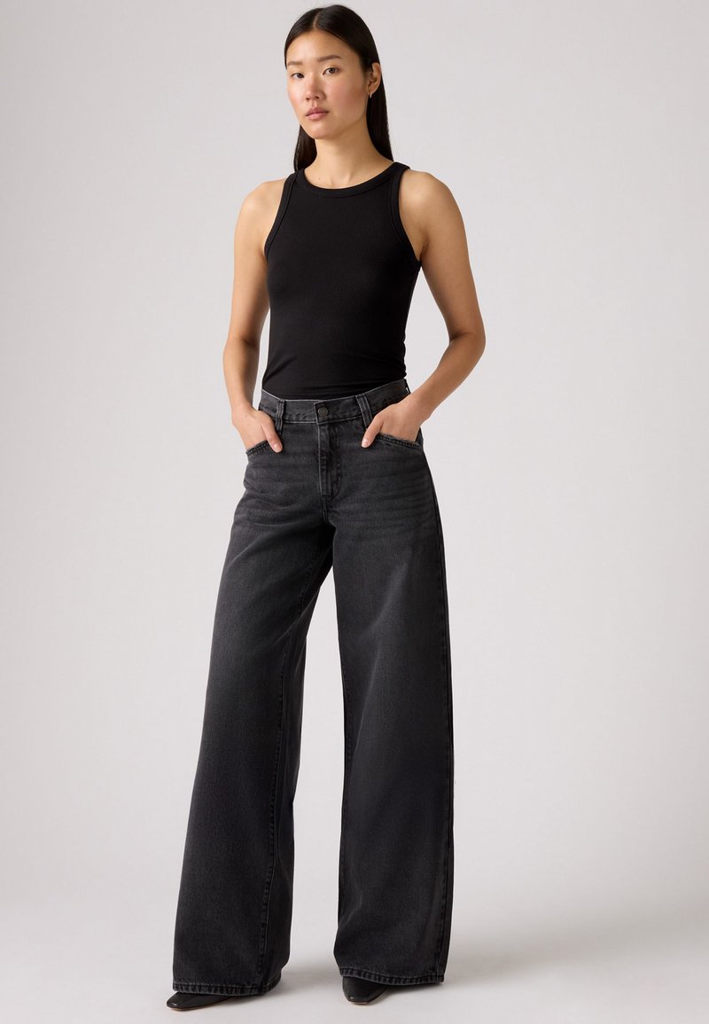 Débardeur noir côtelé associé à un jean taille haute à jambes larges de couleur gris foncé. Le denim comporte des poches avant et une fermeture à boutons.