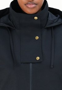 Veste noire avec un col montant, des boutons-pression dorés et une fermeture éclair, fabriquée dans un tissu lisse et léger, avec des cordons de serrage ajustables.