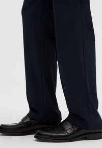 Zapatos de vestir de cuero negro combinados con pantalones de traje azul marino oscuros, contra un fondo claro y liso.