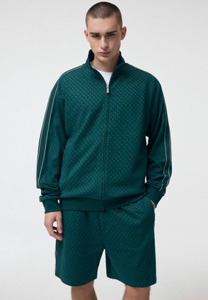 Młody mężczyzna ubrany w ciemnozieloną, wzorzystą kurtkę zapinaną na zamek i pasujące szorty, stojący z rękami w kieszeniach na białym tle.