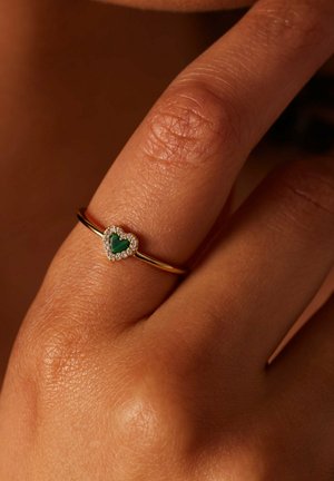 Goldring mit einem herzförmigen grünen Stein, umgeben von kleinen weißen Edelsteinen, getragen an einem Finger mit sichtbarer Hautstruktur.