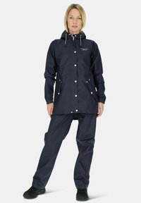 Marineblauwe waterdichte regenjas met een capuchon, drukknoopsluitingen en twee voorzakken, gecombineerd met bijpassende broek en zwarte schoenen.