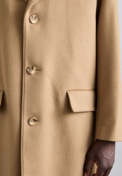 Abrigo de lana beige con cuello de solapa, cinco botones y dos bolsillos frontales, con una textura suave y un ajuste entallado.