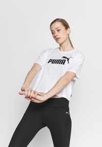 Balta trumpa marškinėliai su juodu "PUMA" logotipu ir siluetu ant krūtinės, derinami su juodomis sportinėmis legingais, turinčiais mažą logotipą.
