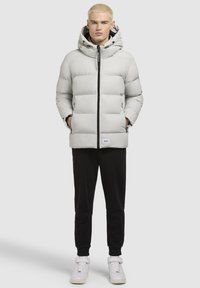 Lys grå dunjakke med hætte, der har skrå syninger, en frontlynlås og sidelommer. Sammen med sorte joggingbukser og hvide sneakers.