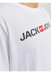 Camiseta de algodón blanca con mangas cortas que presenta el texto en negrita "JACK" y "JONES" en negro, y un acento de logo cuadrado rojo en la parte frontal.