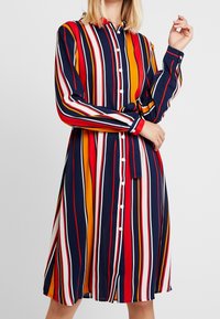 Robe à manches longues à carreaux en bleu marine, rouge, jaune et blanc. Comprend une ceinture et un col. Texture de tissu lisse.