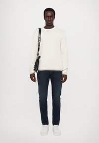 Weißer gerippter Strickpullover, dunkelblaue Jeans, schwarze Umhängetasche und weiße Sneaker. Modell steht vor einem schlichten weißen Hintergrund.