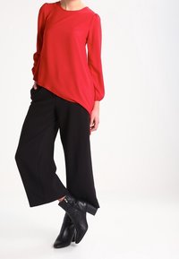 Blusa roja de manga larga con un corte holgado y dobladillo curvado, combinada con pantalones anchos negros y botas negras con tacón cuadrado.