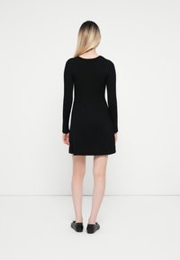 Femme aux cheveux blonds, debout de dos, portant une robe courte noire à manches longues et des chaussures plates noires sur un fond blanc uni.
