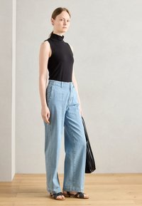 Black sleeveless turtleneck, light blue high-waisted wide-leg jeans, black sandals, and a black tote bag on a wooden floor against a plain wall.