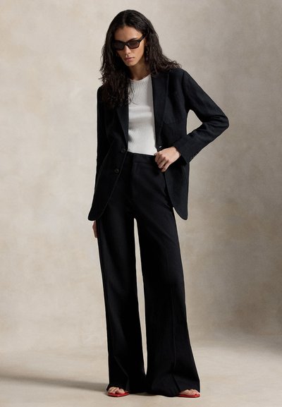 Γυναίκα με μακριά σκούρα μαλλιά που φοράει μαύρα γυαλιά ηλίου, μαύρο σακάκι και παντελόνια με φαρδιά πόδια, λευκό τοπ και κόκκινα σανδάλια με ανοιχτά δάχτυλα, στέκεται μπροστά σε απλό φόντο.