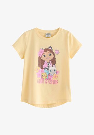 Ung pige med langt hår iført en gul "Gabby & Friends"-trøje, blå jeans og hvide sneakers, gående fremad mod en ensfarvet baggrund.