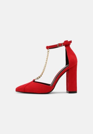 Chaussure à talon haut en daim rouge avec un bout pointu, une bride de cheville réglable et un détail de chaîne en or. Comprend un talon bloc épais.