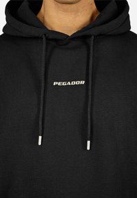 Μαύρη φούτερ μπλούζα με κουκούλα, κατασκευασμένη από βαμβάκι, με ένα μικρό λευκό λογότυπο "PEGADOR". Περιλαμβάνει κορδόνια με ασημί μεταλλικές άκρες.