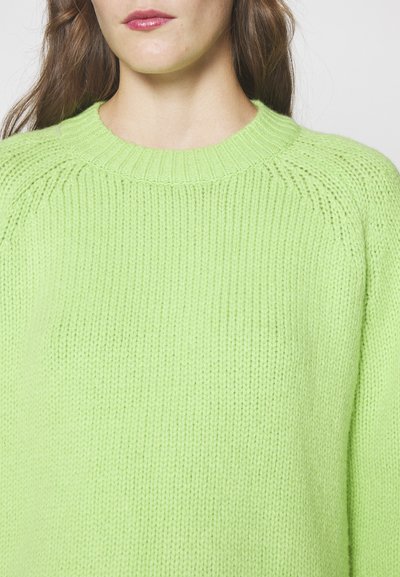 Femme portant un pull en laine vert vif avec un col rond côtelé et des manches longues, montrée de dessous le menton jusqu'au milieu du torse.