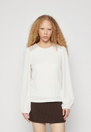 White ribbed long-sleeve top with lace shoulder accents, featuring a rounded neckline and relaxed fit; paired with a striped brown mini skirt.