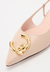 Spiss tå sko i mykt nude skinn med en rosa innersåle, med en stor gullfarget metallakcent i sirkulært design foran.