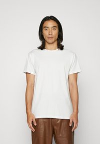 T-shirt blanc en coton à manches courtes avec un col rond, porté par une personne, associé à un pantalon en cuir marron, se tenant devant un fond clair.