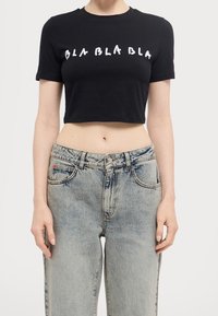 Czarna, krótka koszulka z krótkim rękawem z białym napisem "BLA BLA BLA" nadrukowanym na klatce piersiowej; w połączeniu z jasnoniebieskimi, wysoko osadzonymi dżinsami.