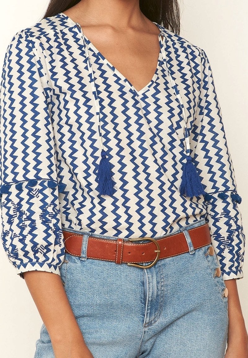Femme portant un chemisier blanc avec un motif zigzag bleu, des pompons au niveau du col, des manches trois-quarts, une ceinture marron et un jean bleu clair avec des boutons sur le côté.