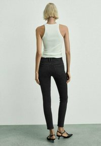 Black skinny jeans with back pockets, paired with a white ribbed tank top and black heeled shoes. Smooth, fitted design, minimal detailing.
