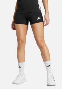 Černé sportovní šortky s těsným střihem, zdobené bílým logem Adidas a akcenty, spárované s bílými crew ponožkami a černými sportovními botami.