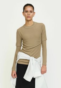 Khaki ribbet kortærmet top med høj hals, parret med en hvid og grå stribet skjorte bundet om livet, og sorte bukser.