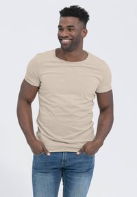 Beżowy t-shirt z krótkim rękawem i okrągłym dekoltem, wykonany z miękkiego materiału. Model nosi niebieskie dżinsy, trzyma ręce w kieszeniach, uśmiechając się na jasnym tle.