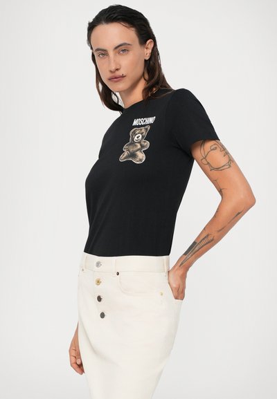 Camiseta negra con gráfico de un oso y texto "Moschino". Llevada con una falda crema de talle alto. Mangas cortas, diseño ajustado, tejido suave.