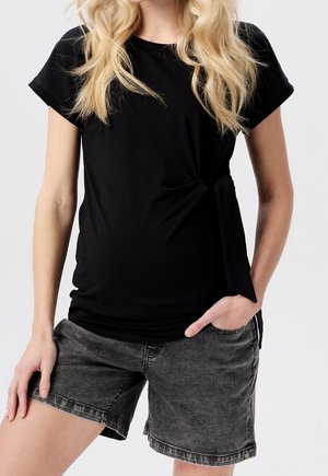 Kobieta o długich blond włosach, ubrane w czarny, zawiązany z boku t-shirt i ciemnoszare krótkie dżinsy, jedna ręka w kieszeni, neutralne tło.