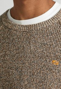 Maglione lavorato a maglia in tonalità marroni e beige con una finitura testurizzata. Presenta un colletto a coste e una piccola etichetta arancione con il marchio sul petto.
