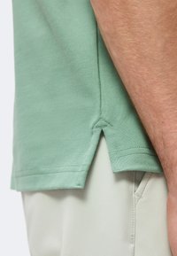 Vue latérale de l'ourlet d'une chemise verte à manches courtes avec une petite fente latérale au-dessus de la poche d'un pantalon beige clair et d'un bras.