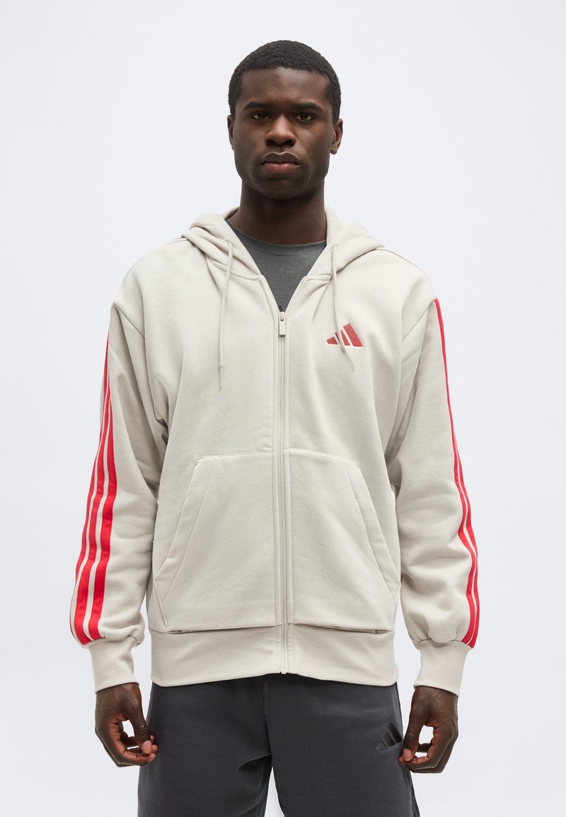 Homme debout portant un sweat à capuche zippé beige clair avec des bandes rouges sur les manches et un pantalon gris foncé avec un logo sur la cuisse gauche.