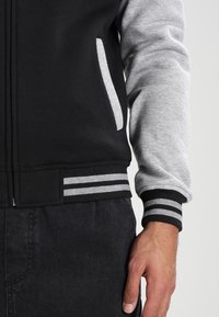 Giacca a zip nera e grigia con maniche grigie, tasche laterali e polsini a righe nere e grigie. Miscela di cotone, texture liscia.