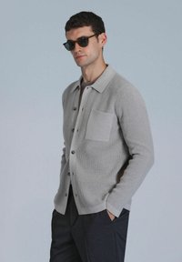 Gray ribbed cardigan with a collared design, featuring a chest pocket and black buttons, worn over a gray shirt and paired with dark pants.