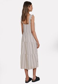 Abito a strisce beige a strati con spalline arricciate e schiena scoperta, abbinato a sandali scuri. Il materiale sembra leggero e informale.