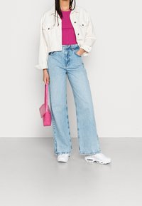 White cropped denim jacket, fuchsia ribbed top, light blue wide-leg jeans, and white sneakers. Holding a pink textured handbag.