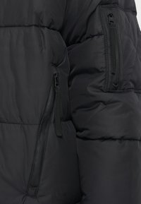 Črna podložena jakna z dvema žepoma na zadrgo. Material se zdi gladek z rahlo sijajno teksturo. Vidna je prešita zasnova.
