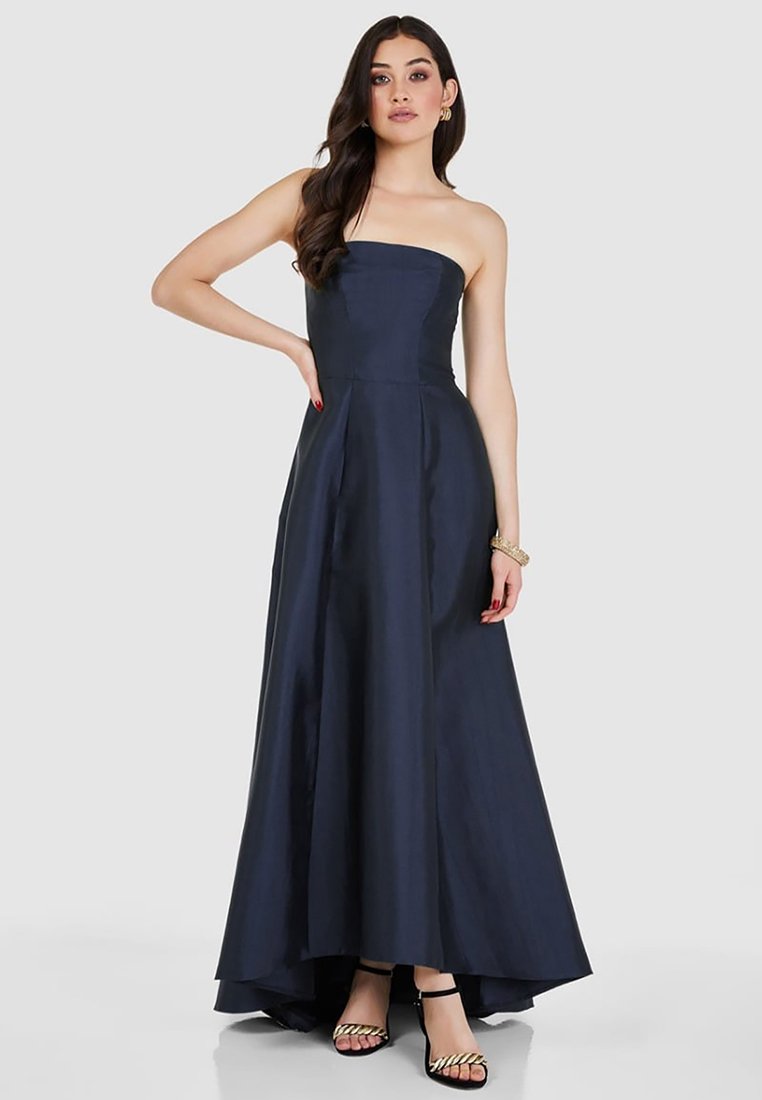 Abito blu navy senza spalline con un corsetto aderente, gonna svasata e orlo asimmetrico. Texture liscia con un leggero lucido. Indossato con sandali neri con cinturini.