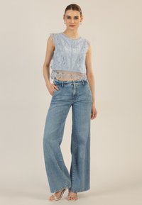 Helles blaues Spitzen-Crop-Top mit gewellten Kanten, kombiniert mit weit geschnittenen hellblauen Jeans. Das Model trägt silberne Riemchensandalen.