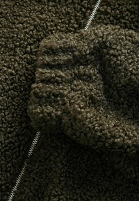 Olivgrüner Fleece-Stoff mit einer weichen, strukturierten Oberfläche und einem sichtbaren Reißverschlussdetail, das entlang des Materials verläuft.