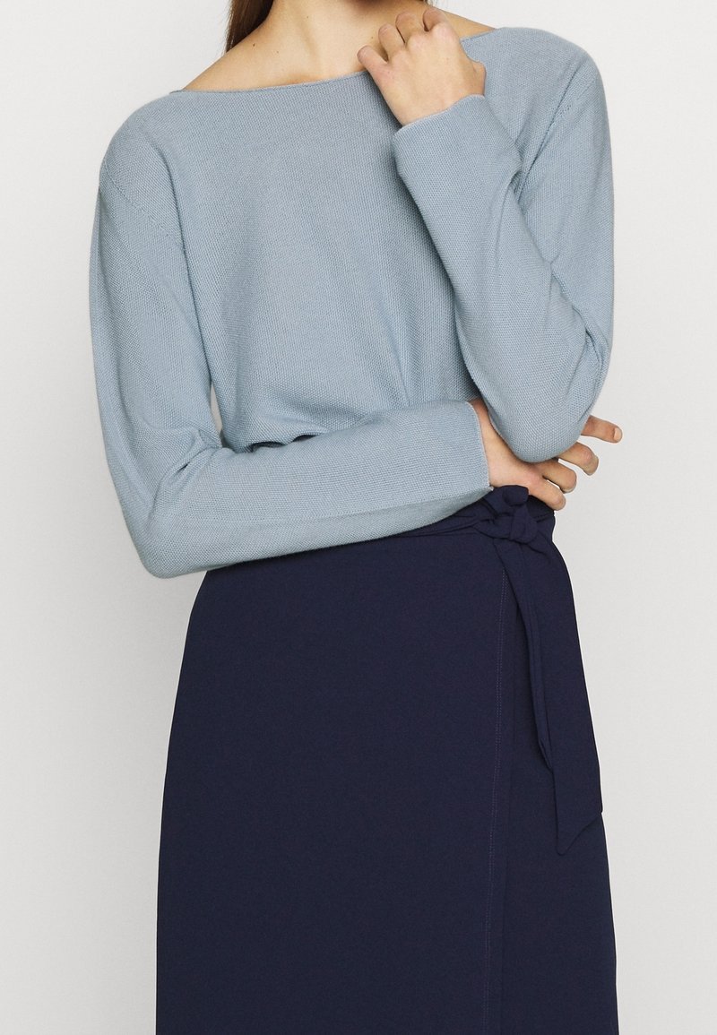 Svetle modrý pletený svetr s dlouhými rukávy a off-shoulder designem. Kombinovaný s temně modrou sukní, která má na pásu detail s vázáním.