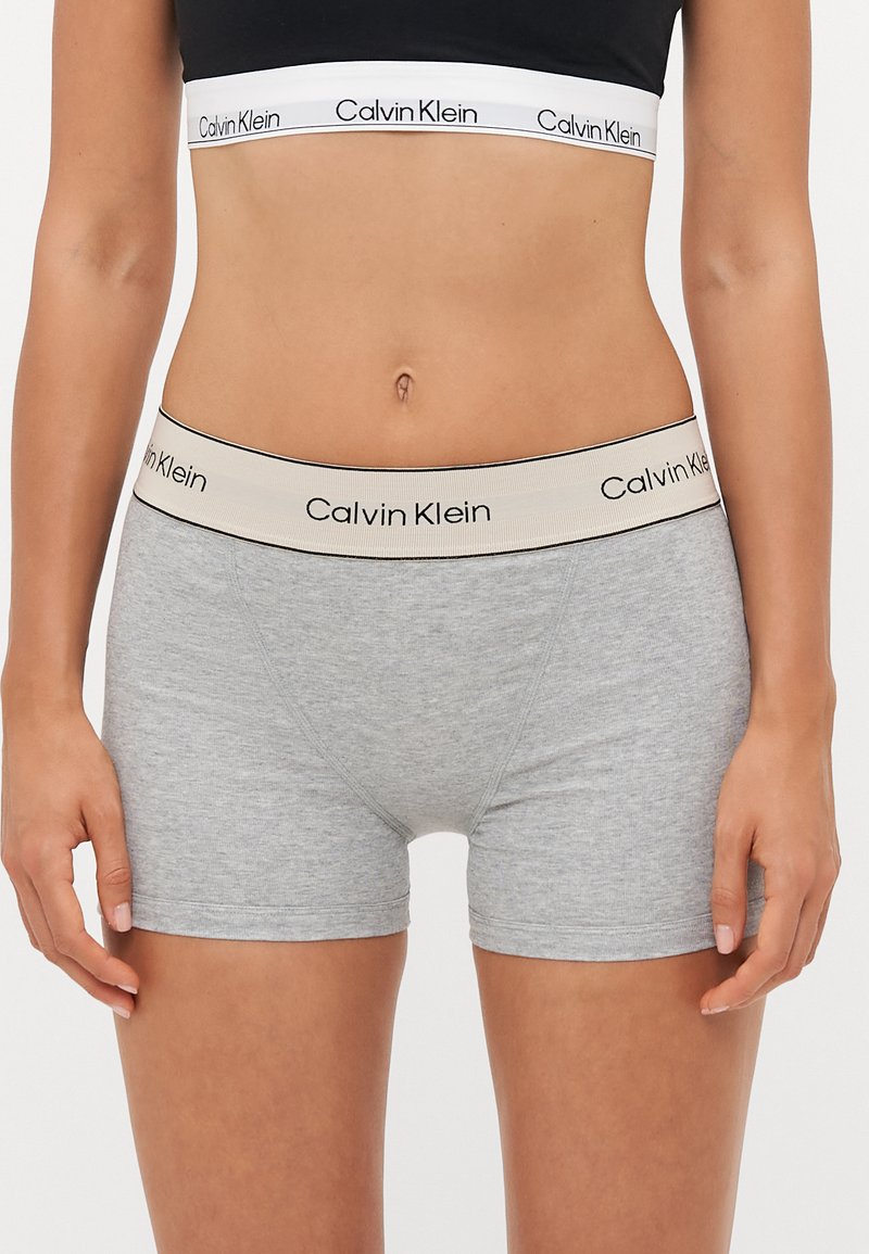 Sive pamučne bokserice s bež elastičnim pojasom na kojem piše "Calvin Klein" u crnoj boji. Bez uzoraka ili dodatnih ukrasa.