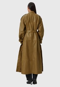 Lange olijfgroene leren trenchcoat met een ceintuur in de taille, grote revers en een gladde textuur. Heeft een split aan de onderkant en opgerolde mouwen.