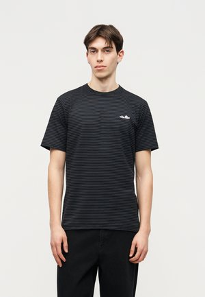 Νέος άνδρας με κοντά σκούρα μαλλιά που φοράει μαύρο και σκούρο γκρι ριγέ μπλουζάκι και μαύρο παντελόνι, στέκεται μπροστά σε απλό ανοιχτό φόντο.