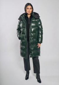Kobieta stojąca, ubrana w długi, błyszczący, ciemnozielony puchowy płaszcz z kapturem, czarny sweter na zamek, ciemne spodnie i czarne botki na obcasie.