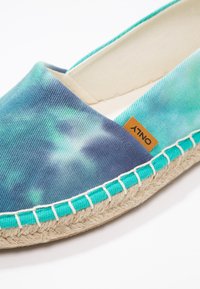 Chaussure slip-on en toile tie-dye bleu et sarcelle avec semelle en jute, présentant une étiquette marron. Couture d'accent turquoise autour du bord de la semelle.