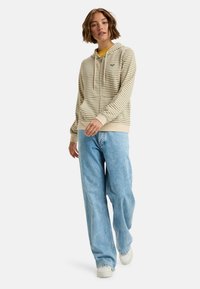 Sudadera beige a rayas con cordones, combinada con jeans de pierna ancha en azul claro. La sudadera cuenta con un pequeño detalle de logo y puños acanalados.