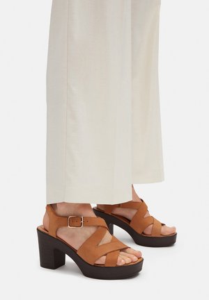 Sandalias de tacón de cuero color cuero con tiras cruzadas, un detalle de hebilla y un tacón plataforma grueso de color marrón oscuro. Textura suave, diseño de punta abierta.